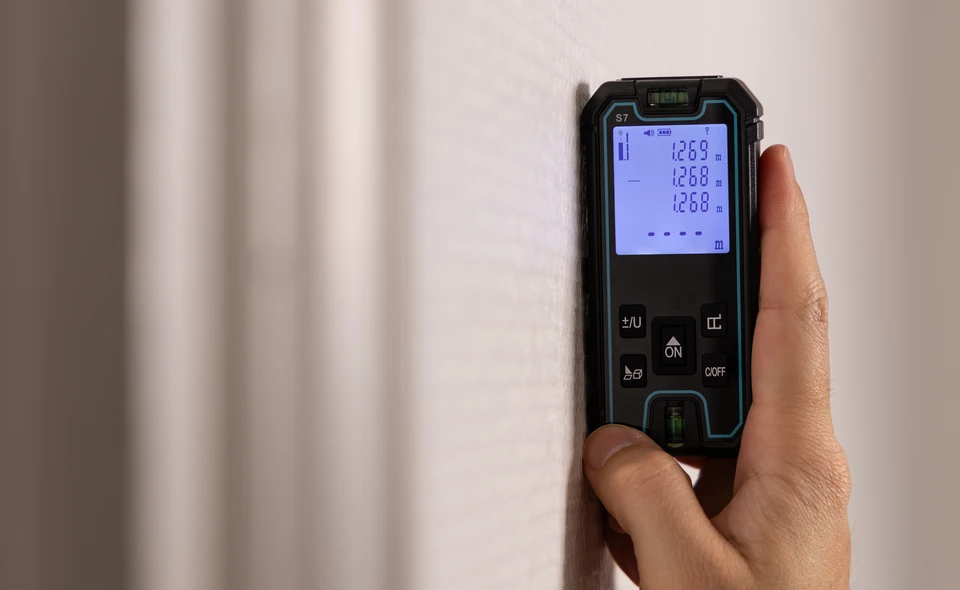 Intérieur d'un logement résidentiel moderne, diagnostiqueur professionnel en tenue civile mesurant la longueur d'une pièce avec un décamètre laser ou un mètre ruban, dans une chambre ou un salon lumineux. L'image montre le processus de mesurage rigoureux et méthodique de la surface habitable d'un bien immobilier.